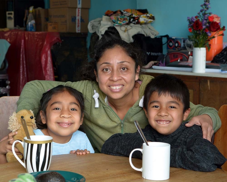 Astrid sits at a table inside the family house with her brother. Behind them, Lissette kneels and wraps her arms around them. They all share a smile.