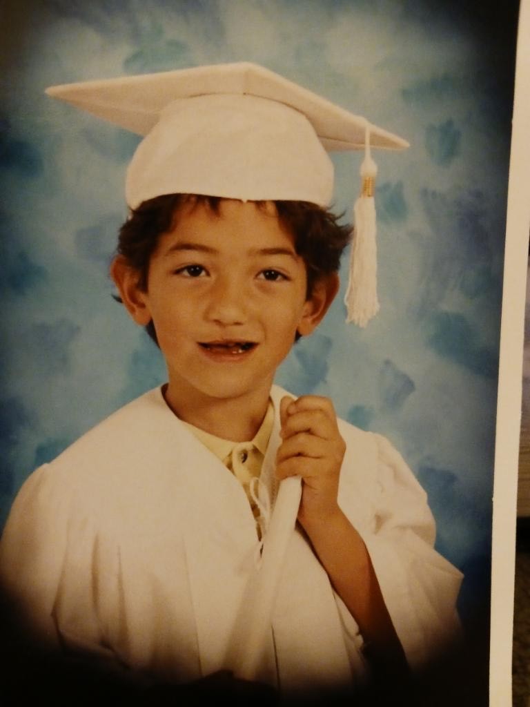 Young Dillon posing for a graduation photo in a white cap and gown holding his diploma